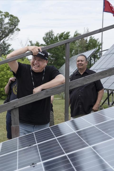 Cody Two Bears smiles while leaning on a solar panel array — energy sovereignty