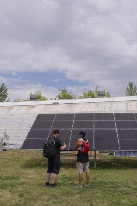 Charlee Rising sun stands in front of a solar array speaking to another expert. 