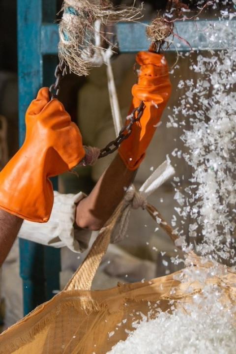 A worker at a plastic recycling facility wears orange gloves to empty small pieces of plastic into a large bin — plastic credits
