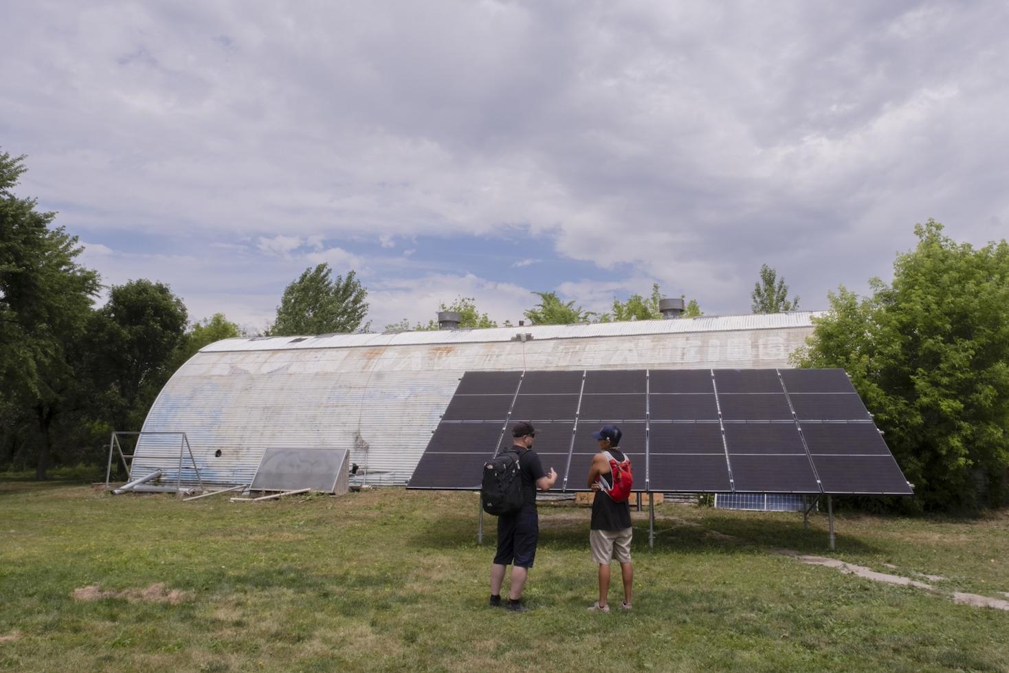 Charlee Rising sun stands in front of a solar array speaking to another expert. 