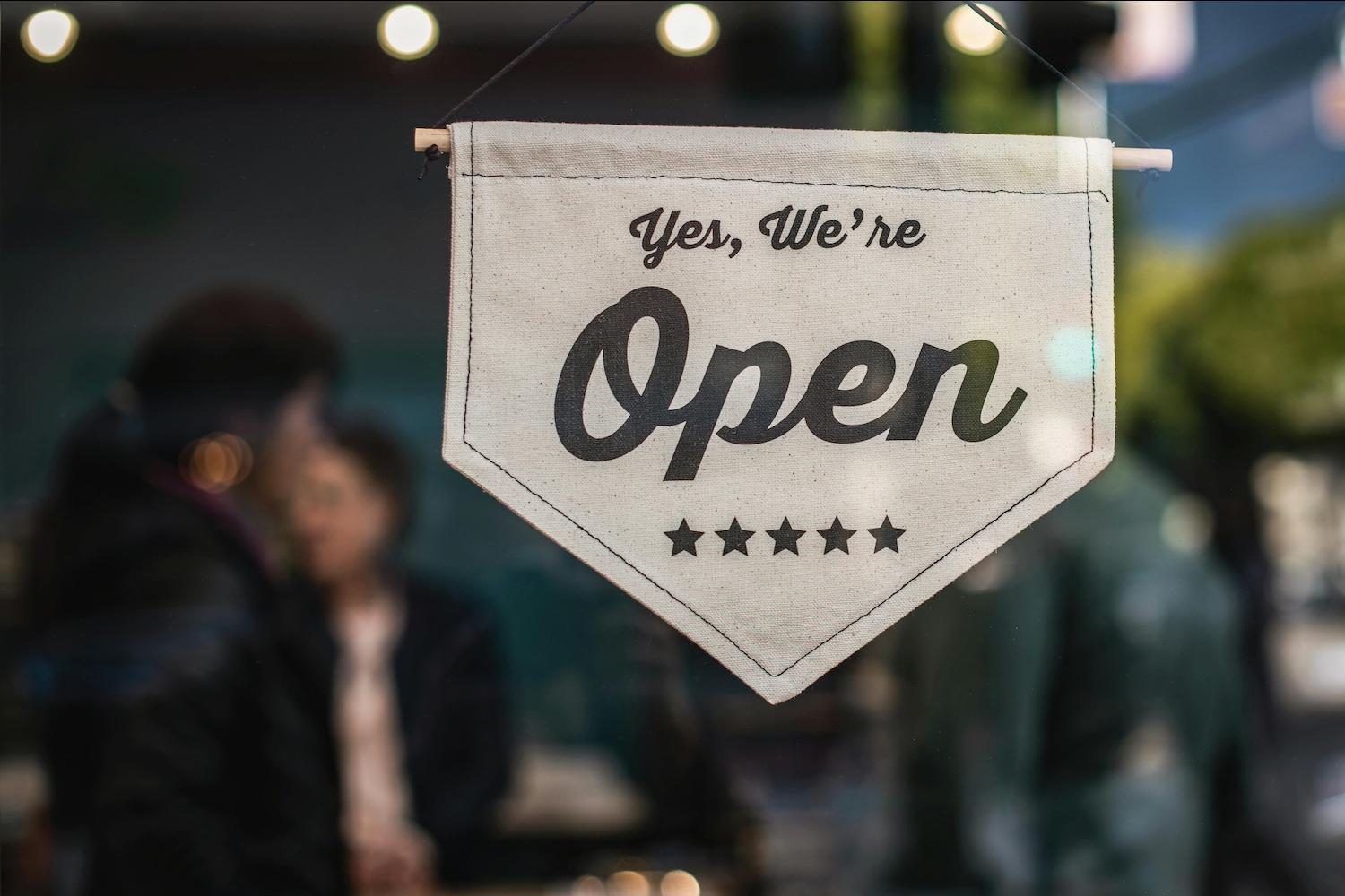 A sign on a window reads "yes, we're open" — social impact 