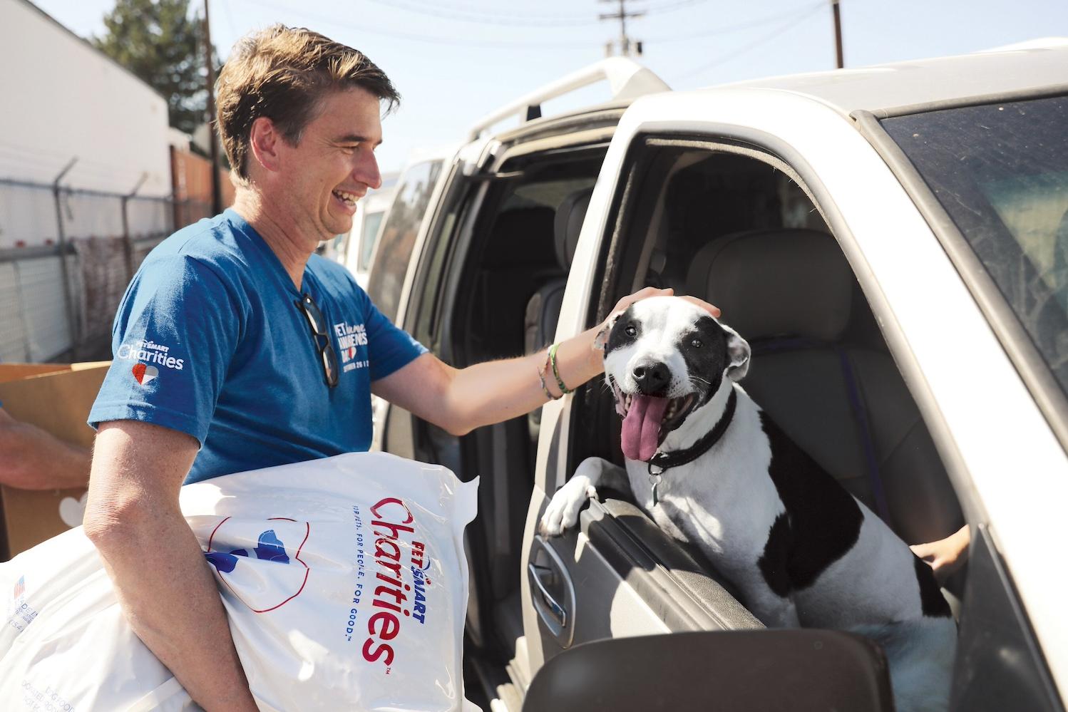 A PetSmart Charities volunteer petting a dog in car and holding a bag of dog food — food insecurity