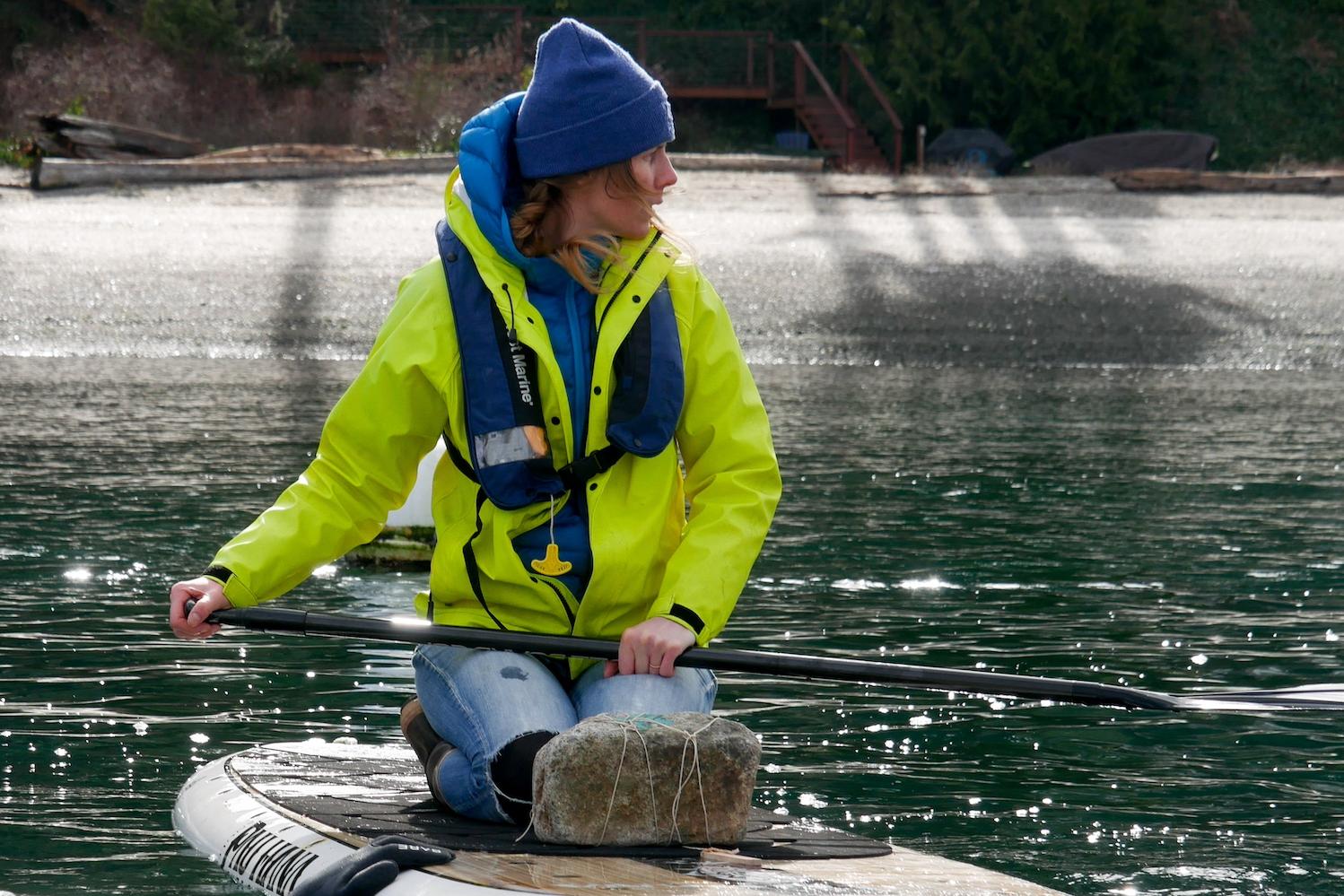 A member of the Puget Sound Restoration Fund team paddle boards in the water carrying a rock with string attached for bull kelp gardening.