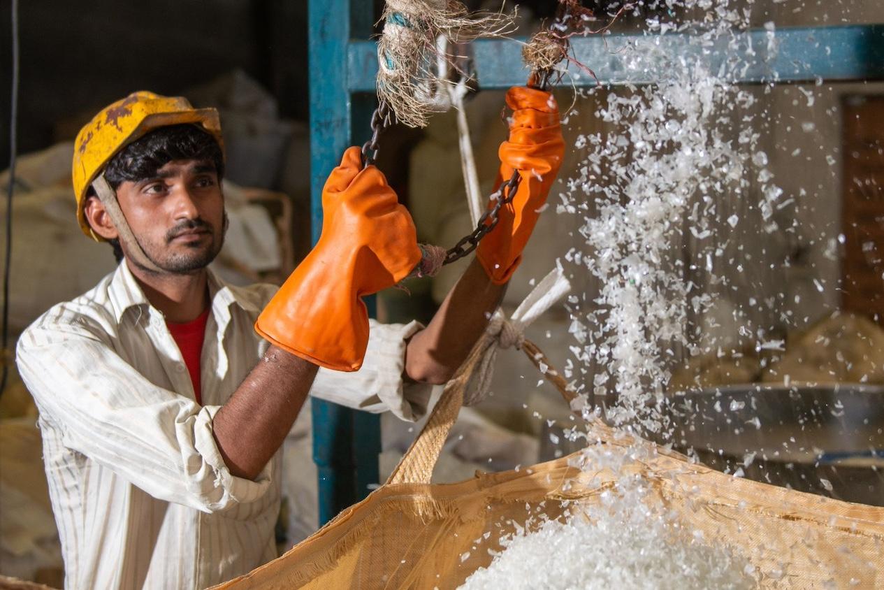 A worker at a plastic recycling facility wears orange gloves to empty small pieces of plastic into a large bin — plastic credits