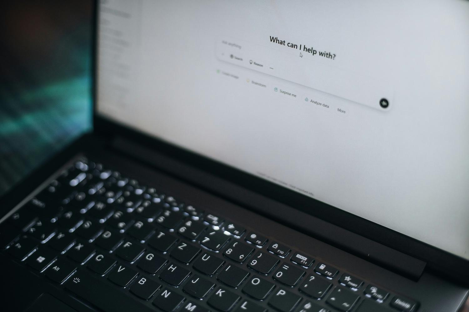 An open laptop displays the ChatGPT homepage, which reads, "What can I help with?" — AI